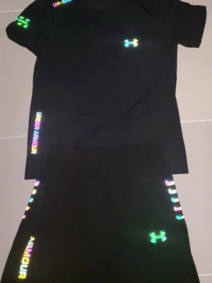 Svart träningsset från Under Armour - Snyggt svart träningsset från Under Armour med t-shirt och shorts. Setet har reflekterande loggor och text i regnbågsfärger på både tröja och byxa. Tillverkat i ett lätt och snabbtorkande material som passar perfekt till träning eller sport. Passar också S