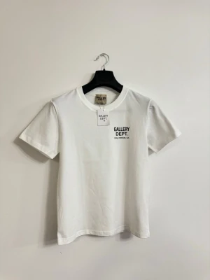 Vit t-shirt  Gallery Dept. - Säljer en clean vit t-shirt från Gallery Dept. med svart tryck på bröstet där det står 'GALLERY DEPT. HOLLYWOOD, CA.'. Klassisk rund hals och korta ärmar. Perfekt basic-plagg för en enkel och snygg stil.