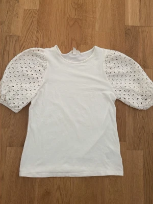 Vit topp med broderade puffärmar - Snygg vit topp med puffiga, broderade ärmar i spets och rund halsringning. Kroppen är i mjuk bomull och ärmarna har ett vackert hålmönster som ger en romantisk vibe. Perfekt att styla med jeans eller kjol för en trendig look.