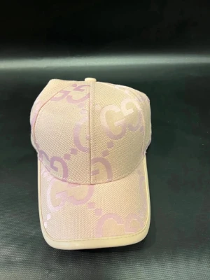 Beige och rosa keps från Gucci - Snygg beige keps från Gucci med ljusrosa GG-mönster över hela kepsen. Justerbar rem bak i ljust läder med guldfärgad detalj. Tillverkad i Italien och har en klassisk böjd skärm. Perfekt accessoar för en trendig look.
