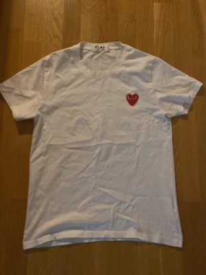 Vit t-shirt med rött hjärta PLAY - Vit t-shirt med rund hals och korta ärmar. På bröstet sitter ett litet rött hjärta med ögon, typiskt för PLAY-kollektionen. Enkel och clean design som passar till det mesta. Tillverkad i mjuk bomull för skön känsla.