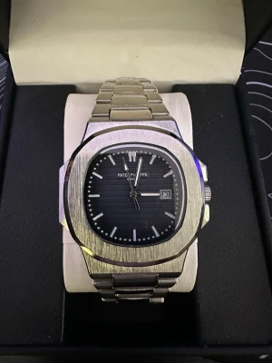 Patek  - Stilren PP Nautilus klocka i borstat silverstål med svart urtavla och datumvisning. Klockan har en ikonisk rundad fyrkantig form och länkarmband i metall. Perfekt för dig som gillar exklusiv design och klassiska detaljer.
