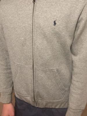 Grå zip hoodie från Polo Ralph Lauren - Snygg gråmelerad hoodie från Polo Ralph Lauren med dragkedja framtill och två fickor. Klassisk mörkblå logga broderad på bröstet. Tillverkad i mjuk bomullsblandning och har ribbade muddar vid ärmslut och nederkant.