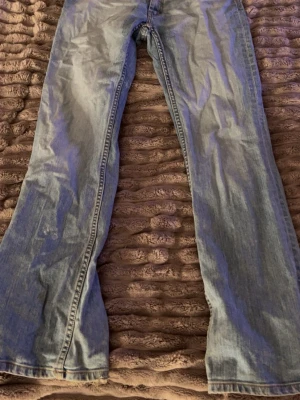 Ljusblå bootcut jeans - Säljer ett par ljusblå bootcut jeans med slitningar och klassisk femficksmodell. Jeansen har en snygg tvättad look och stängs med knapp och dragkedja. Perfekta för dig som gillar retrovibbar och avslappnad stil.