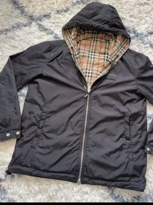 Svart burberry windbreaker  - Svart vindjacka med dragkedja och huva. Insidan av huvan har ett klassiskt rutigt mönster i beige, svart och rött. Jackan har två fickor framtill och knappar vid ärmsluten. Perfekt för en stilren och avslappnad look.