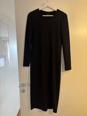 Svart långärmad långklänning - En enkel och stilren svart långklänning med rund halsringning och långa ärmar. Klänningen har en rak passform och är tillverkad i ett mjukt, ribbat material som ger en bekväm känsla. Perfekt för dig som gillar minimalistisk stil.