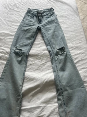Ljusblå bootcut jeans med hål - Säljer ett par ljusblå bootcut jeans med slitna hål på båda knäna. Jeansen har klassisk femficksmodell och är tillverkade i ett mjukt denimtyg. Perfekta för dig som gillar en avslappnad och trendig stil.