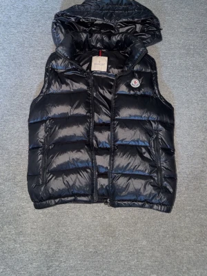 Moncler väst - Snygg svart dunväst från Moncler med glansig finish och avtagbar huva. Västen har två fickor med dragkedja och klassisk Moncler-logga på bröstet. Perfekt för lager på lager och ger en trendig look till din outfit.