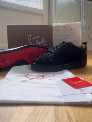 Christian Louboutin svarta sneakers - Säljer ett par svarta Christian Louboutin sneakers med klassisk röd sula och svarta nitar på tån. Skorna har lågt skaft, snörning och är tillverkade i läder. Kommer med originalkartong, dustbag och certifikat.