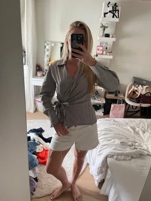 Randig omlottblus med knytband - Snygg omlottblus med smala vita och grå ränder. Blusen har långa ärmar, klassisk krage och knytband i midjan för en feminin siluett. Tillverkad i ett lätt tyg som känns skönt mot huden. Perfekt att styla med jeans eller kjol.