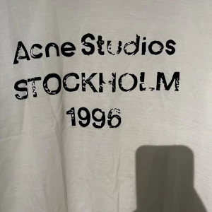 Vit Acne Studios t-shirt Stockholm - Vit t-shirt från Acne Studios med svart tryck framtill där det står 'Acne Studios STOCKHOLM 1996'. Klassisk passform och rund hals. Perfekt för dig som gillar stilrena och trendiga plagg.