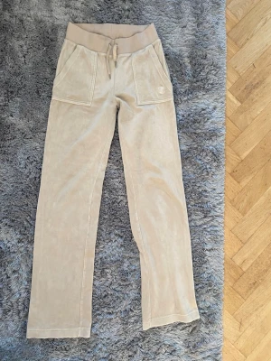  Juicy Couture  - Säljer ett par beige mjukisbyxor i mjukt velourmaterial med bred resårmidja och dragsko. Byxorna har raka ben och två stora fickor framtill. Perfekta för en avslappnad och chill stil.
