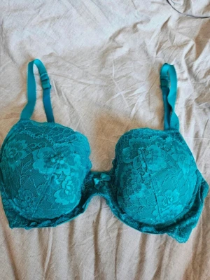 BH  H&M 80D - Snygg turkos bh med vadderade kupor och vacker spets med blommönster. Justerbara axelband och liten rosett framtill. Bh:n har hakknäppning bak och ger bra stöd. Perfekt för dig som vill ha något både bekvämt och stilrent.