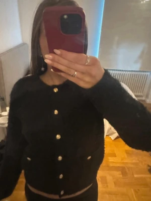 Svart fluffig kofta med guldknappar - Snygg svart kofta i fluffigt material med dekorativa guldknappar framtill. Koftan har två fickor fram och en rak, något kortare passform. Perfekt att styla till jeans eller kjol för en trendig look. 💗💗