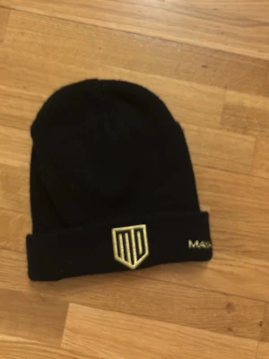 Svart mössa från Maya Delorez - Snygg svart mössa från Maya Delorez med uppvikt kant. Framtill finns en guldfärgad MD-logga och broderad text i guld. Perfekt accessoar för kalla dagar och passar till de flesta outfits.