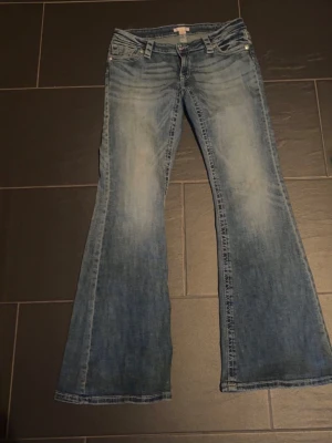 Blå bootcut jeans med slitningar - Säljer ett par blå bootcut jeans med snygga slitningar och ljus tvätt. Jeansen har klassisk femficksdesign, låg midja och vida ben. Perfekta för dig som gillar retrostil och vill ha ett par jeans som sticker ut.  Jeans ifrån gina 14+ aldrig använda så väldigt bra skick säljer då de är för stora
