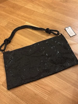 Svart glittrig kuvertväska Stories - Snygg svart kuvertväska från Stories med glittrande mesh och ett flätat axelband. Väskan har rektangulär form och stängs med dragkedja. Perfekt för att lyfta din outfit med lite extra bling.
