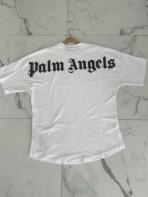 Vit Palm Angels t-shirt - Säljer en vit t-shirt från Palm Angels med stor svart logga i gotisk stil på ryggen och mindre logga vid halsen fram. Klassisk passform och rund halsringning. Perfekt för dig som gillar streetwear och vill sticka ut.