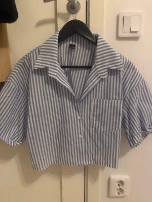Blårandig kortärmad skjorta - Säljer en blå- och vitrandig skjorta med korta ärmar och en avslappnad, boxig passform. Skjortan har en klassisk krage, knäppning framtill och en bröstficka. Materialet ser ut att vara bomull, perfekt för en chill och trendig look.