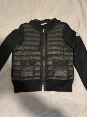 Moncler cardigan med dun – svart – storlek XS/S (storlek 2)  - Säljer min fina Moncler-cardigan i svart med dun framtill och stickad ull på ärmar och rygg. En klassisk och stilren modell som passar perfekt till höst, vinter och vår.  Cardiganen är i mycket fint skick med endast normalt slitage. Inga hål eller större skador. Alla detaljer, knappar, dragkedja, märken och etiketter finns kvar.  * Storlek: 2 (motsvarar ungefär XS) * Färg: Svart * Original QR-kod och etiketter finns kvar * Från rökfritt hem  Jag har tyvärr inget kvitto, därför har jag lagt upp t