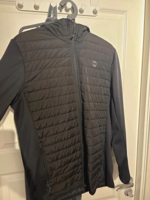 Svart quiltad vindjacka med huva - Snygg svart vindjacka med quiltat mönster framtill och på huvan. Jackan har hel dragkedja, två sidofickor med dragkedja och en liten logga på bröstet. Ärmar och sidor är i ett mjukare material för extra komfort.