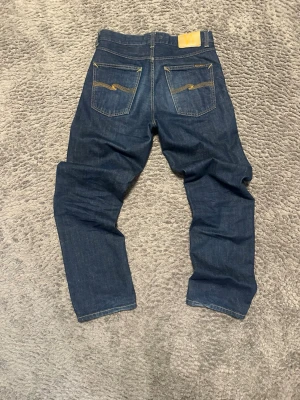 Rad Rufus nudie jeans  - Säljer ett par mörkblå jeans från Nudie Jeans med raka ben och klassisk femficksmodell. Jeansen har kontrastsömmar och snygga broderade detaljer på bakfickorna. Tillverkade i slitstark denim som ger en tidlös look.
