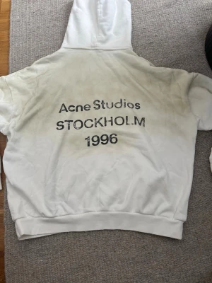 Vit hoodie Acne Studios Stockholm - Säljer en vit hoodie från Acne Studios med tryck på ryggen där det står 'Acne Studios STOCKHOLM 1996' i svart text. Hoodien har klassisk passform, huva och ribbade muddar. Perfekt för dig som gillar streetwear och enkel design.