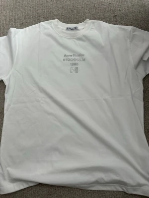 Vit t-shirt från Acne Studios - Säljer en vit t-shirt från Acne Studios med glittrig text och logga på bröstet. T-shirten har klassisk passform och rund hals. Perfekt för dig som gillar stilrena plagg med en twist.