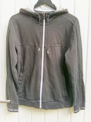 HUGO BOSS hoodie med dragkedja - Snygg mörkgrå hoodie från HUGO BOSS med vit dragkedja och justerbar huva med snören. BOSS-logga på bröstet och tryck längs huva och ärmslut. Tillverkad i mjukt bomullsmaterial, perfekt för en avslappnad streetwear-look.