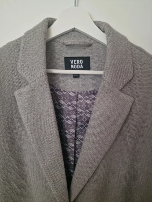 Grå kappa från Vero Moda - Stilren grå kappa från Vero Moda med klassisk krage, en knapp framtill och två stora fickor. Kappan har ett mönstrat innerfoder och rak passform som ger en snygg och modern look. Perfekt för dig som vill ha en enkel men trendig ytterjacka.