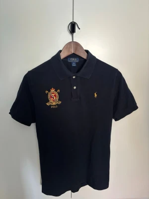 Svart pikétröja från Ralph Lauren - Klassisk svart pikétröja från Ralph Lauren med gul broderad logga på bröstet och en stor brodyr med siffran 3 och texten POLO. Tröjan har korta ärmar, krage och två knappar vid halsen. Perfekt för en stilren och sportig look.