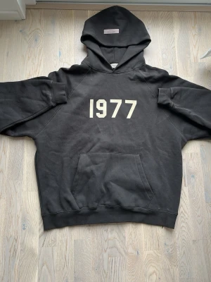 Svart Essentials hoodie med 1977-tryck - Säljer en svart hoodie från Essentials med stort vitt 1977-tryck på bröstet. Hoodien har en klassisk känguruficka framtill och en rejäl huva. Tillverkad i mjukt bomullsmaterial som är perfekt för en avslappnad streetwear-look.