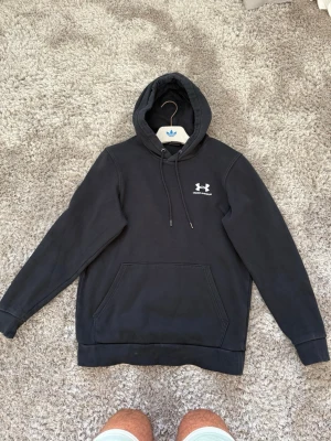 Svart hoodie från Under Armour - Säljer en svart hoodie från Under Armour med klassisk logga på bröstet. Hoodien har huva med dragsko och en stor magficka framtill. Perfekt för dig som gillar sportig och enkel stil. Materialet känns mjukt och bekvämt.