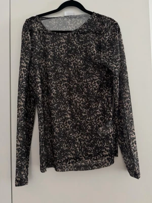 Svart mesh topp med abstrakt mönster - Snygg långärmad mesh topp i svart med abstrakt mönster. Toppen är genomskinlig och har en rund halsringning. Perfekt att styla lager på lager för en trendig look. 