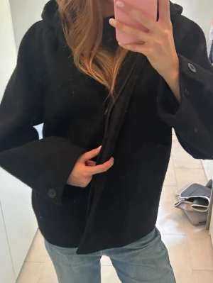 Svart vid kappa med huva - Säljer en svart kappa med  huva. Kappan har stora ärmar med knappdetaljer vid ärmsluten och stängs framtill. Perfekt för kyligare dagar och enkel att styla till olika outfits.