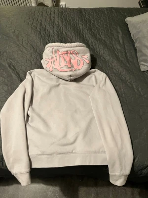 Ljusgrå och rosa (dress) - Säljer en ljusgrå hoodie med rosa snören och coolt rosa graffiti-tryck på huvan. Hoodien har en klassisk känguruficka och ribbade muddar. Perfekt för dig som gillar streetstyle och vill ha något bekvämt och snyggt.