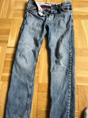 Blå jeans från Gant - Snygga blå jeans från gant med klassisk femficksdesign och raka ben. Jeansen har detaljer i rött och vitt vid fickorna och linningen samt en liten logga på fickan. Perfekta för en avslappnad och trendig stil.
