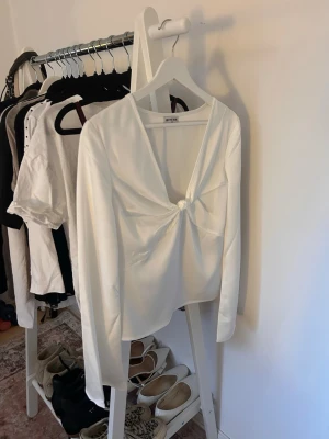 Vit långärmad blus med knut -  vit silkmaterialsliknande blus från MEET ME THERE i storlek S/M. Blusen har lång ärm, djup v-ringning och en knutdetalj framtill. 