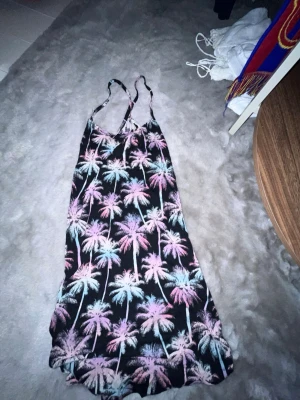 Palmmönstrad långklänning H&M - Säljer en svart långklänning från H&M med färgglatt palmmönster i rosa, blått och vitt. Klänningen har smala axelband och är perfekt för sommardagar. Luftigt material och avslappnad passform.