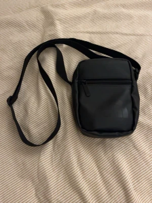 Svart axelväska från The North Face - Snygg svart axelväska från The North Face med justerbar rem och dragkedja framtill. Väskan har en stilren design med diskret logga på framsidan och är tillverkad i ett tåligt, vattenavvisande material. Perfekt för att bära det viktigaste på stan eller resan.