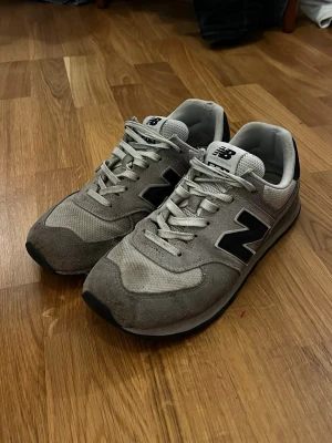 New Balance 574 - Snygga grå sneakers från New Balance med klassisk svart N-logga på sidan. Skorna har ovandel i mocka och mesh, vita skosnören och en bekväm sula. Perfekta för dig som gillar stilrena och sportiga skor.