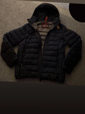 S Svart dunjacka med huva från Parajumpers  - Säljer en svart dunjacka från Parajumpers med huva och dragkedja. Jackan har quiltad design, två sidofickor med dragkedja och detaljer i orange. Insidan är ljusgrå och jackan har klassiska,  två små hål i jackan. Parajumpers-loggor på bröstet och insidan.
