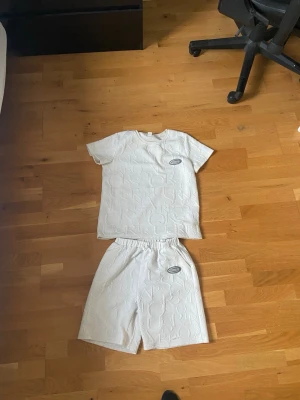 Vitt set med strukturmönster - Säljer ett vitt set med t-shirt och shorts i mjukt material. Båda delarna har ett upphöjt strukturmönster och en liten oval patch framtill. T-shirten har rund hals och korta ärmar, shortsen har resår i midjan för skön passform.