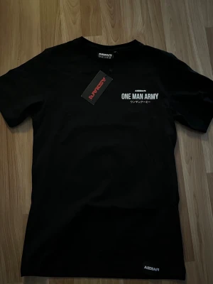 ASKARI T-shirt  - Svart t-shirt från ASKARI med trycket 'ONE MAN ARMY' och japanska tecken på bröstet. Klassisk rund hals och korta ärmar. Perfekt för dig som gillar streetwear och vill sticka ut med ett coolt statement. Aldrig använd bara testad.