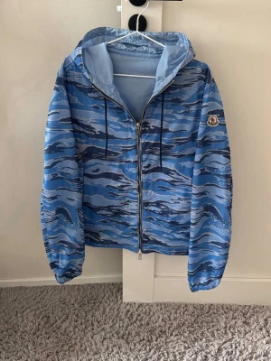 Moncler wind jacket - Snygg vändbar vindjacka från Moncler i ljusblått på ena sidan och blått kamouflagemönster på andra. Jackan har huva, dragkedja framtill och Moncler-logga på ärmen. Tillverkad i lätt, vindtätt material och har elastiska muddar vid ärmslut.