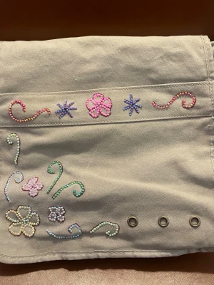 Beige ryggsäck med pärlbroderi - Säljer en beige ryggsäck i tyg med justerbar axelrem. Väskan har färgglada pärlbroderier i form av blommor och snirklar på framsidan samt metallöljetter längst ner. Perfekt för dig som gillar en unik och kreativ stil!