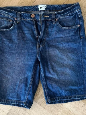 Mörkblå jeansshorts med fem fickor - Snygga mörkblå jeansshorts med klassisk femficksdesign och kontrastsömmar. Shortsen har knapp och dragkedja framtill samt bälteshällor. Perfekta för varma dagar och passar till det mesta i garderoben.