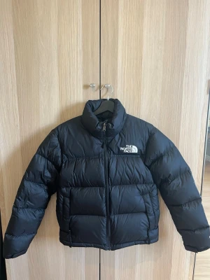 Svart pufferjacka från The North Face - Säljer en svart pufferjacka från The North Face med hög krage och dragkedja framtill. Jackan har två stora fickor på sidorna och logga broderad i vitt på både bröstet och ryggen. Den är endast använd 2/3 gånger så inprincip ny. Storlek: S funkar för både män och kvinnor. Köpt för över 2000kr 