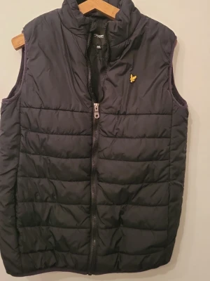 Svart dunväst från Lyle & Scott - Snygg svart dunväst från Lyle & Scott med gul broderad logga på bröstet. Västen har hög krage, dragkedja framtill och quiltad design. Perfekt för lager-på-lager och håller dig varm under kyliga dagar.