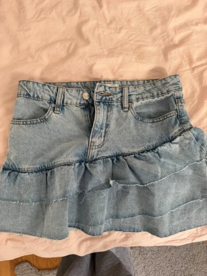 Blå volangkjol i denim - Säljer en kort jeanskjol i ljusblå denim med volangdetaljer och råa kanter. Kjolen har klassiska fickor fram och bak samt bälteshällor. Perfekt för dig som gillar en avslappnad och trendig stil.